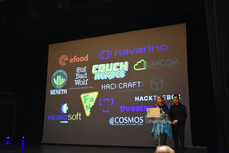 Two presenters at a podium on stage in front of a slide of sponsor logos